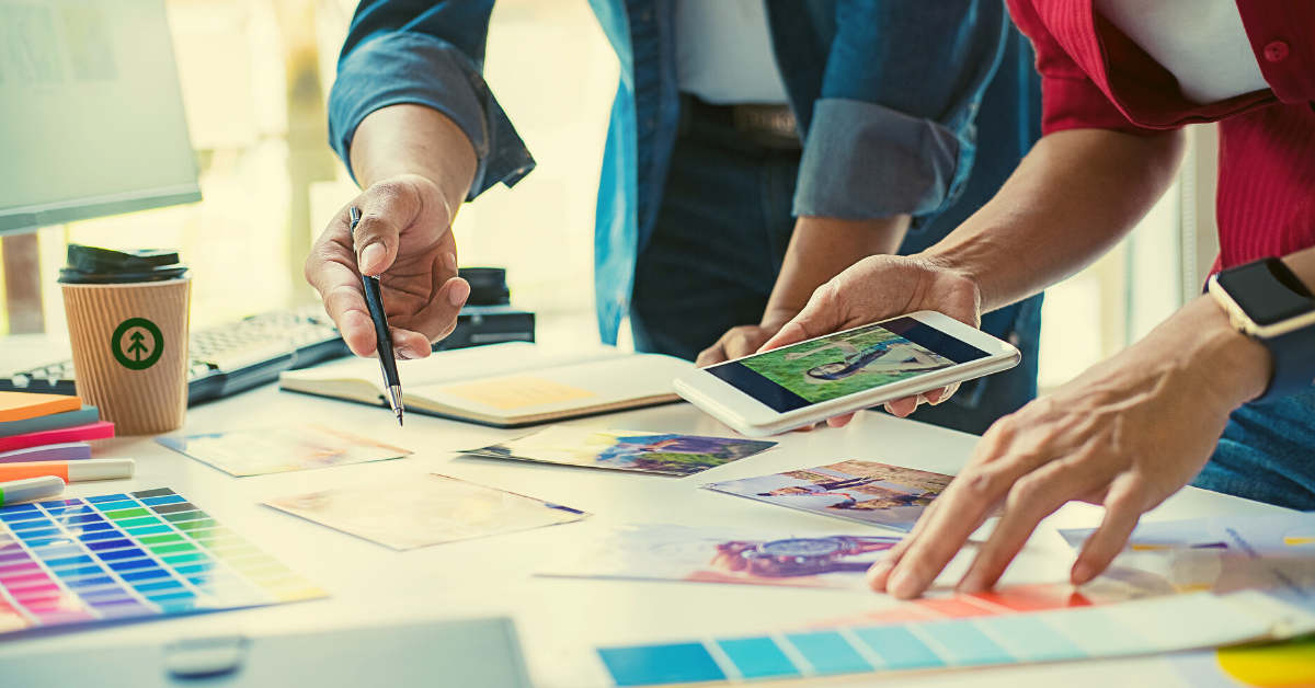 Two website designers looking at a mobile device and choosing colors and images for a website design project from pictures and color swatches laying on a table.