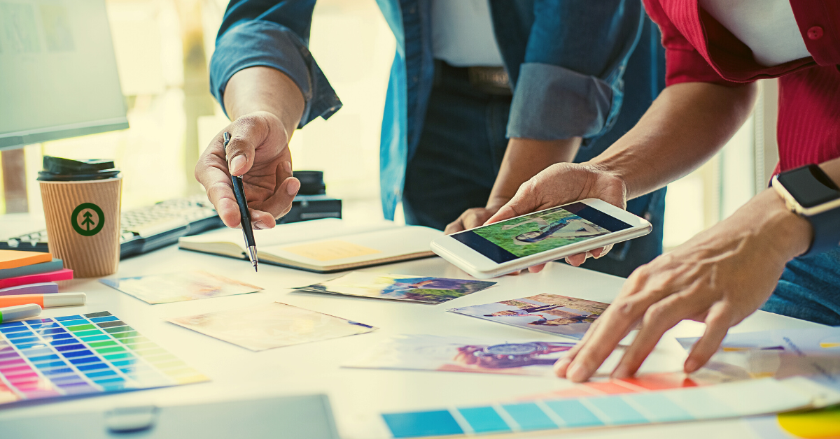 Two website designers looking at a mobile device and choosing colors and images for a website design project from pictures and color swatches laying on a table. 