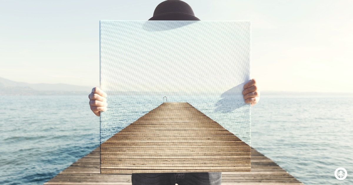 A person standing at the end of a pier over a placid lake holding a painting of the same pier so that it covers his face and makes it appear that his body is missing. Just his hat, legs, and hands are showing.