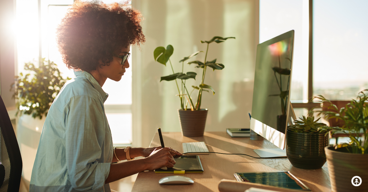 A graphic designer working remotely on a desktop computer in a home office with plants and natural light. 