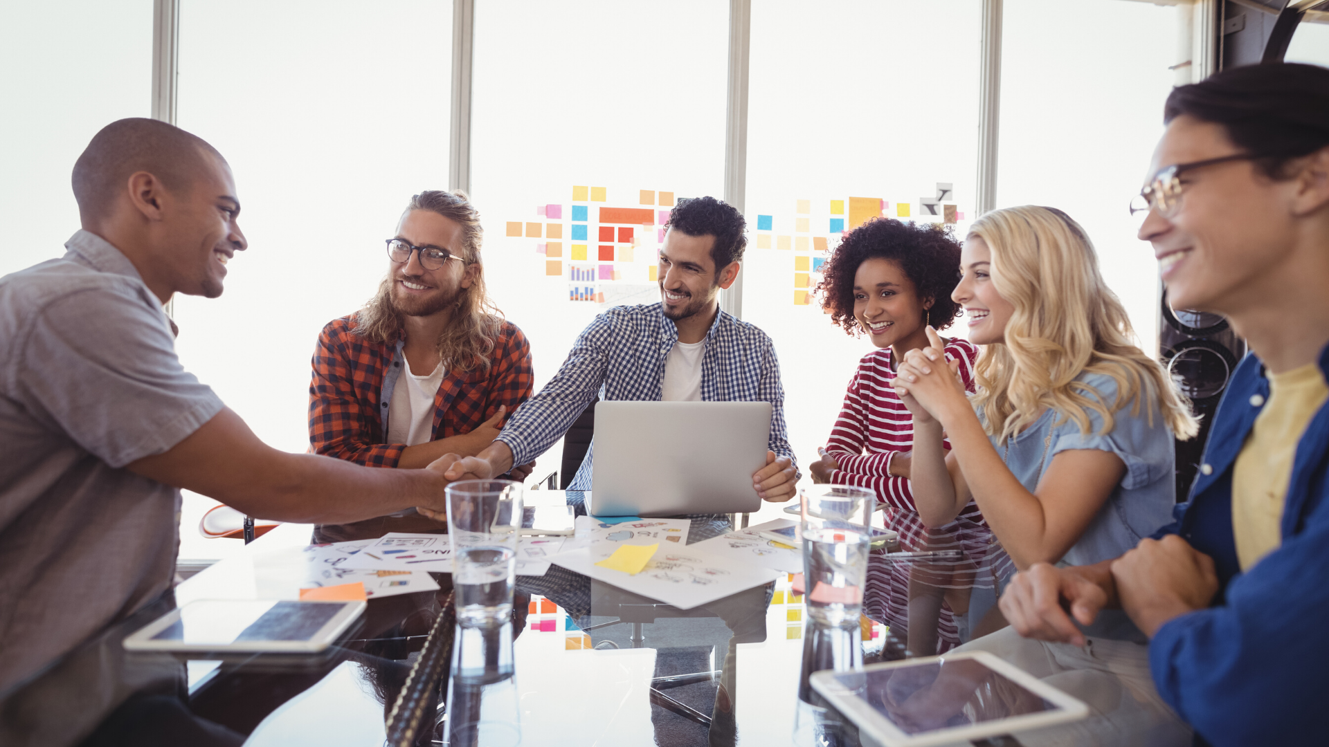 An excited group of coworkers smiling and shaking hands with a  new partner in a tech office. 