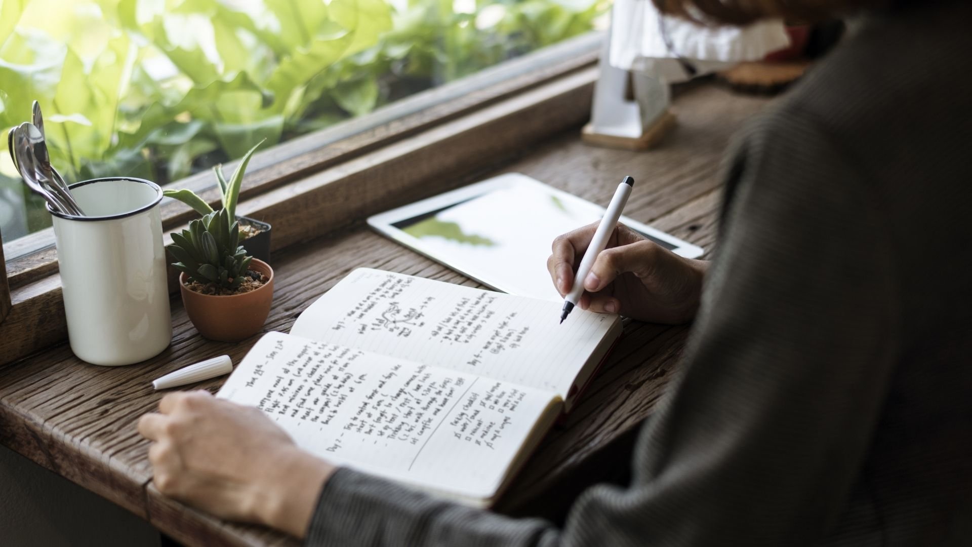 A person writing with a pen in a journal on a table near a green window.