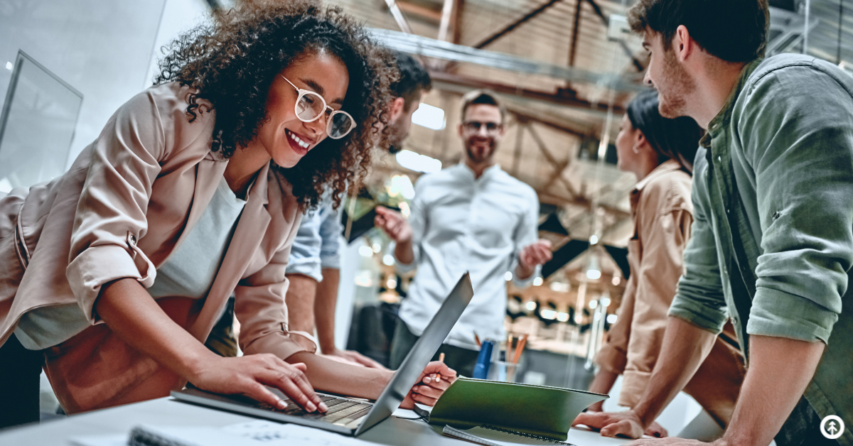 A person with glasses typing on a laptop while surrounded by an excited team of other business people in a professional setting. 